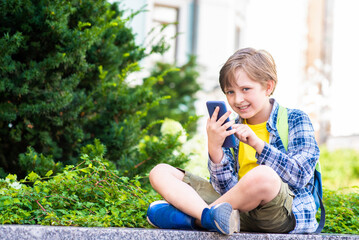 happy handsome boy with backpack sits in the park after school, calls parents or friends, communicates by video call, using mobile outside