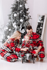 Fototapeta na wymiar Young happy family in the same pyjamas: smiling mom, dad are having fun with baby boy on toy retro car by the decorative Christmas tree. The theme of the family holiday is New year and Xmas.