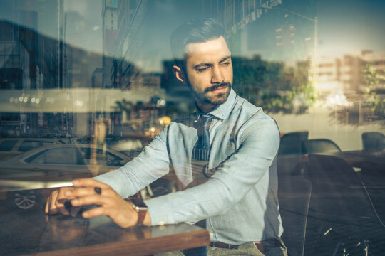 Portrait of handsome business man sitting in a cafe and looking through the window with reflections of urban city.
