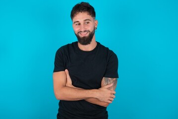 Dreamy rest relaxed young hispanic bearded man wearing black T-shirt standing against blue background crossing arms, looks good copyspace