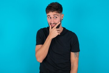 young hispanic bearded man wearing black T-shirt standing against blue background Thinking worried about a question, concerned and nervous with hand on chin.