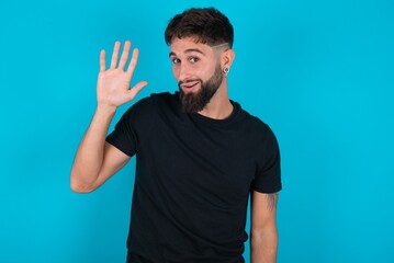 young hispanic bearded man wearing black T-shirt standing against blue background Waiving saying hello happy and smiling, friendly welcome gesture.