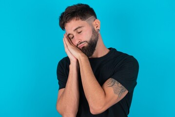 young hispanic bearded man wearing black T-shirt standing against blue background sleeping tired dreaming and posing with hands together while smiling with closed eyes.