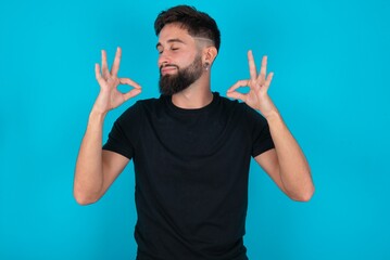 young hispanic bearded man wearing black T-shirt standing against blue background relax and smiling with eyes closed doing meditation gesture with fingers. Yoga concept.