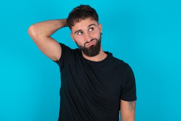 young bearded hispanic man wearing black T-shirt over blue background confuse and wonder about question. Uncertain with doubt, thinking with hand on head. Pensive concept.