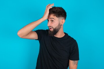 young bearded hispanic man wearing black T-shirt over blue background surprised with hand on head...