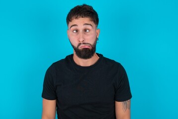 young bearded hispanic man wearing black T-shirt over blue background making grimace and crazy...