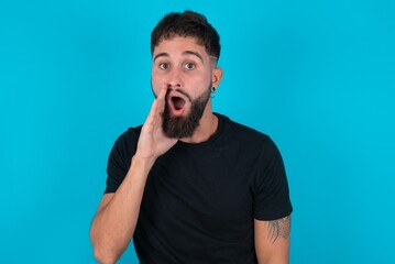 Shocked young bearded hispanic man wearing black T-shirt over blue background looks with great surprisment being very stunned, astonished with unexpected news, Facial expressions concept.