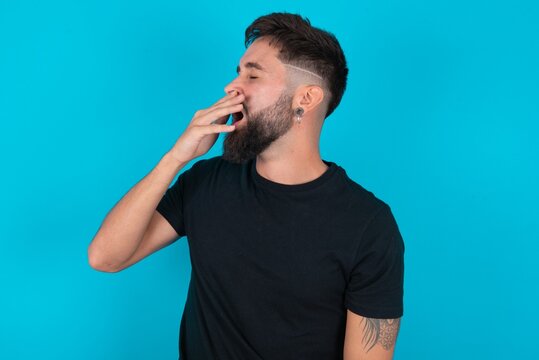 Young Bearded Hispanic Man Wearing Black T-shirt Over Blue Background Being Tired And Yawning After Spending All Day At Work.