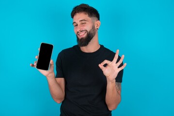young bearded hispanic man wearing black T-shirt over blue background holding in hands cell showing...