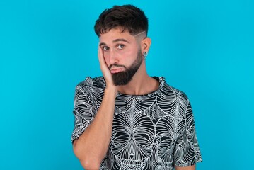 Sad lonely young bearded hispanic man wearing casual clothes over blue background touches cheek with hand bites lower lip and gazes with displeasure. Bad emotions