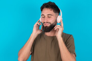 Pleased young bearded hispanic man wearing casual clothes over blue background enjoys listening pleasant melody keeps hands on stereo headphones closes eyes. Spending free time with music