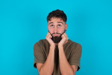 Portrait of sad young bearded hispanic man wearing casual clothes over blue background hands face look empty space