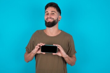 Cheerful cheery content young handsome latin man wearing casual clothes standing over blue background holding in hands device hobby smm post blog