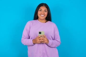 Smiling young beautiful latin woman wearing casual clothes standing against blue background friendly and happily holding mobile phone taking selfie in mirror.