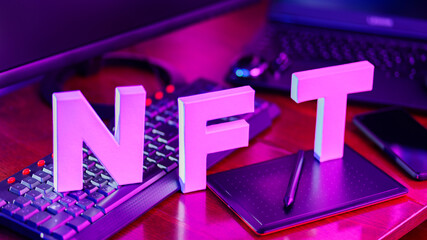 NFT inscription made from 3D letters printed on a 3D printer. NFT lettering at workplace with graphics tablet, smartphone and computer equipment. Non-fungible token in neon pink-blue light