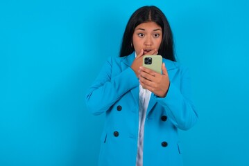 young hispanic businesswoman wearing blue blazer over blue background being deeply surprised,...
