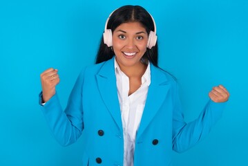 Emotional Young latin businesswoman wearing blue b exclaims loudly feels like winner raises clenched fists keeps mouth opened wears stereo headphones on ears makes yes gesture, listens favourite music