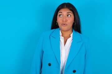 Shocked young latin businesswoman wearing blue blazer over blue background look empty space with open mouth screaming: Oh My God! I can't believe this.