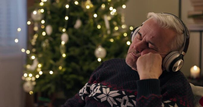 Winter evening. A senior man sits by Christmas tree and listens to relaxing music in headphones. The man dressed in warm holiday sweater, rested head on hand, closed eyes and fell asleep