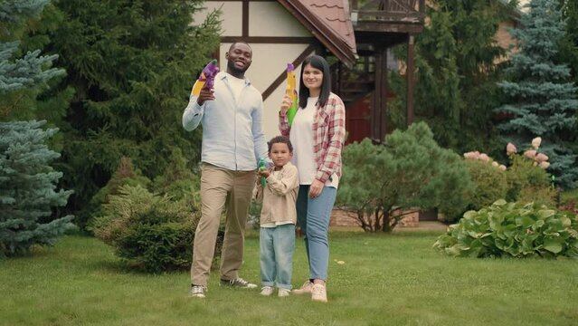 Portrait of a cheerful family with water guns in their hands near the house on the lawn. African dad, Caucasian mom, son.Multiracial Family,Mixed Race,Diverse People,Multiethnic Relations