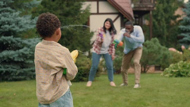 Cheerful family shoot at each other with water guns near the house on the lawn. African dad, Caucasian mom, son.Multiracial Family,Mixed Race,Diverse People,Multiethnic Relations
