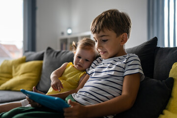 Fototapeta premium One boy sitting with little sister at home looking at digital table