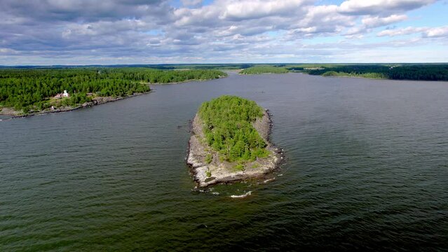 Flying over a rocky island with a few tourists and pine trees in Lake Vanern in Varmland Sweden. On the coast a stuga in the traditional Falu red color