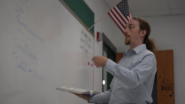 Male school teacher holding book teaching english in school classroom at a whiteboard, with US flag.