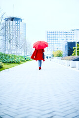 Executive woman walking under a red umbrella in a rainy day, she is dressed in blue jeans and red coat looking to the side. Background of an office building. Copy space Business executive woman.