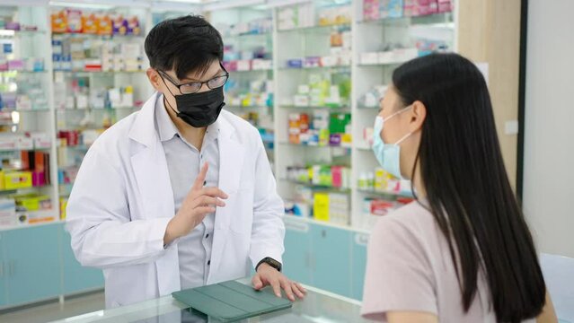 Asian man pharmacist wear surgical face mask medication recommendation about medicine, drugs and supplements to woman patient customer in drugstore. Medical pharmacy and healthcare providers concept.