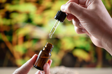 Hands holding a bottle of CBD oil and its dropper lid, with hemp leaf in the background. Legalized CBD product for medical purposes.
