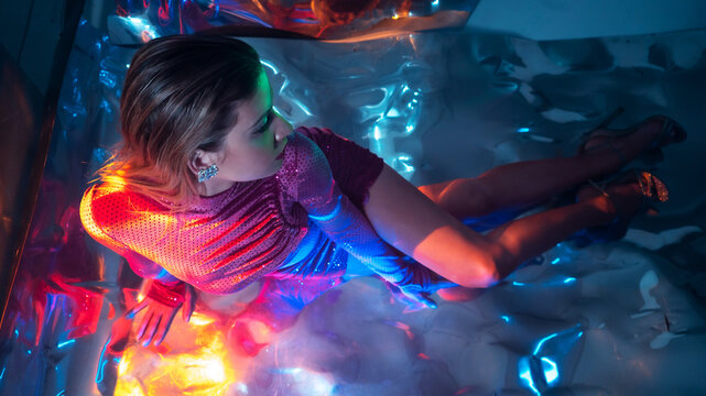 Glamorous Portrait Of A Young Beautiful Blonde In A Stylish Scarlet Short Dress, Sitting On The Floor, Refraction Effects Of Colored Light, Futuristic Fashion Shooting In Neon Light