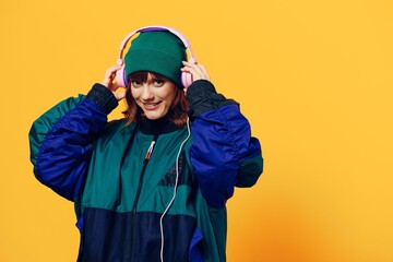 a happy, joyful woman in a green jacket and hat enjoys music standing in pink headphones on a yellow background holding them with her hands and looking at the camera