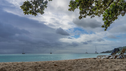 Green tree branches hang over the beach. Two chaise lounges stand on the sand. Yachts are visible on the calm turquoise ocean. Clouds in the blue sky. Seychelles. Mahe Island. Beau Vallon