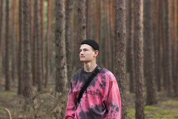 Thoughtful young man in casual clothing and black hat with shoulder belt bag walking in a pine autumn forest.