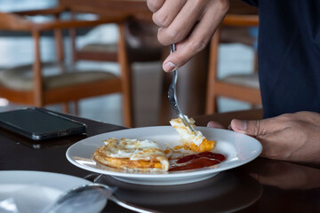Cropped image of a young casual man eating fried eggs with sauce for breakfast. A mobile phone on the side.