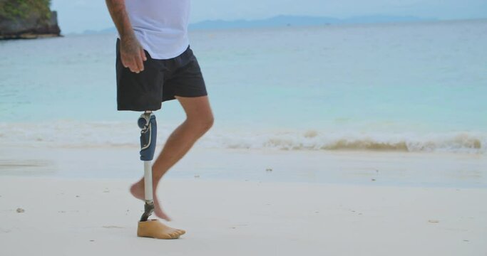 Unknown man with artificial bionic legs prosthesis walking along sandy beach in summer. Unrecognizable disability male spending summer holiday at seaside in slow motion. Concept of tourism and vacatio