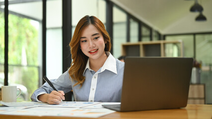 Young employee searching information on laptop and making notes on notebook