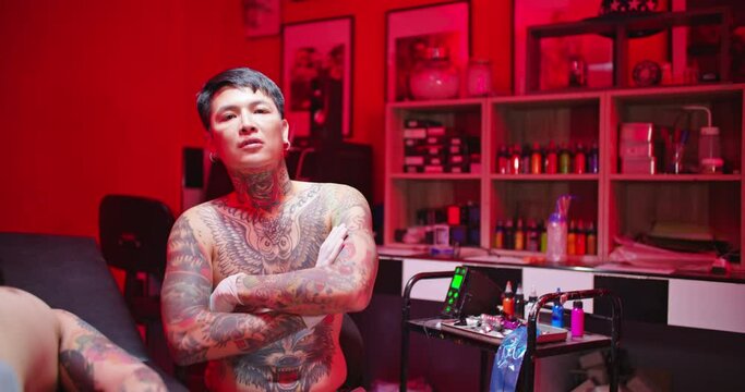Portrait shot of professional man tattooist in dark red room. Young asian male tattoo artist looking at camera with confident in beauty parlor studio. Slow motion. Alternative lifestyle, art.