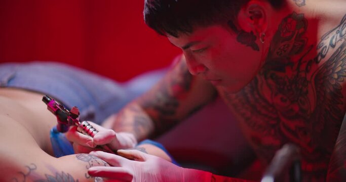 Professional man tattooist tattooing on customers arm in dark room. Close-up young asian male tattoo artist in gloves works with electric rotary needle machine on clients body in beauty parlor studio.