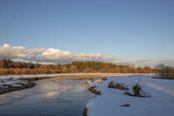 March sunny evening by the river. Blue sky over the horizon. A picturesque landscape, early spring, a river with snow-covered banks, dry grass and bushes. The first thaws, the snow is melting