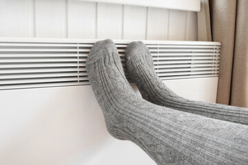 Legs in knitted socks warming over electric heater. Frozen woman wearing a warm woolen socks freezing for winter cold. Discomfort spending time at home. Girl warming her feet on modern radiator