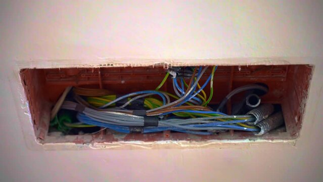 Opened junction box with colourful electricity wires inside the wall on construction site, stack of cables wrapped with black insulting tape, electricity works at home