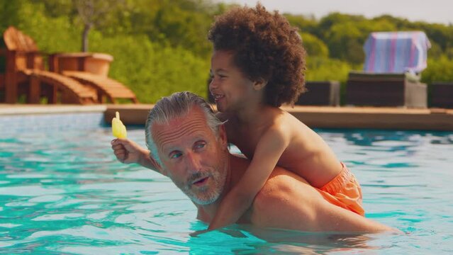 Grandfather playing with grandson eating ice lolly in swimming pool on family summer holiday - shot in slow motion
