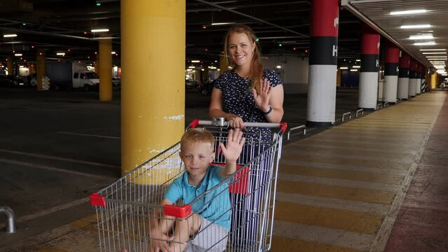 A mother and child are having fun with a cart in the parking lot of a large hypermarket, they wave and look at the camera. Portrait of a boy in a grocery cart and his happy mother.