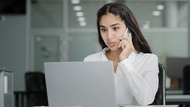 Young indian woman sits in office talk on phone looks at screen laptop make order in online store orders delivery businesswoman consultant remote advises client on telephone using computer application