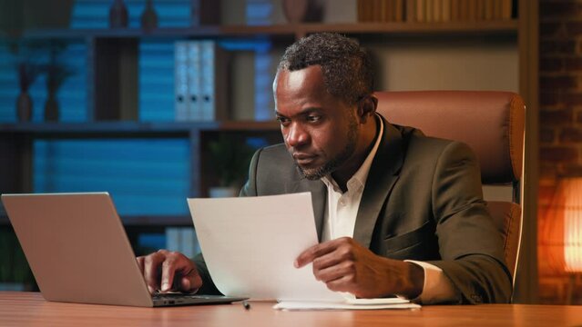 Serious focused businessman sits in office browsing paper document financial report entering information in laptop copying text in electronic form comparing documents developing project analyzes data