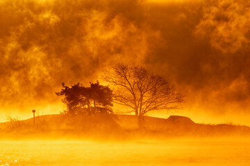 Flaming morning sunrise behind rocky island and  two trees on an extremely cold winter morning at sunrise with sea fog & dramatic clouds and icy Baltic sea on the background in Helsinki, Finland.