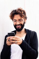smiling young man with beard in a white wall typing happy on mobile phone, concept of urban lifestyle and technology of communication